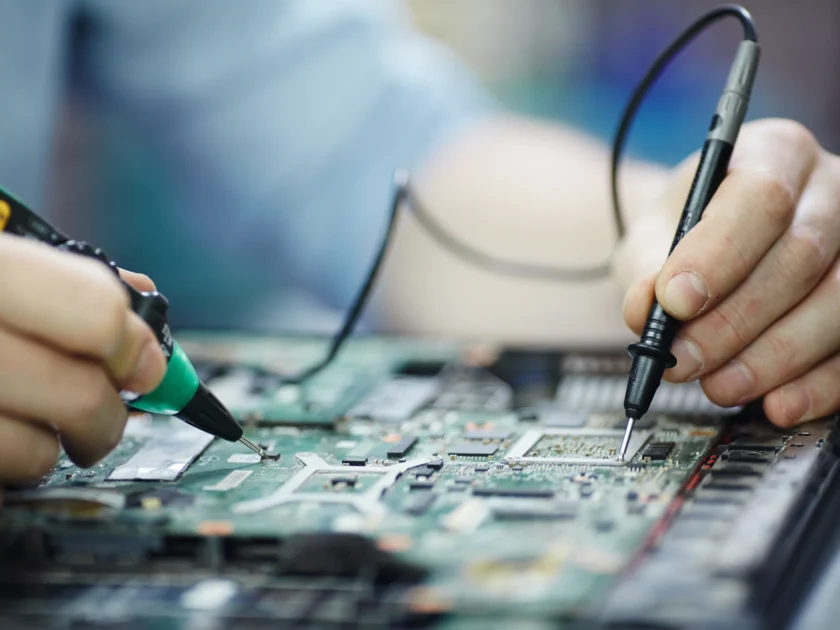 Gros plan sur les mains d’un technicien utilisant deux sondes de multimètre pour tester les connexions et vérifier le courant sur une carte mère d’ordinateur portable. L’image illustre un processus de diagnostic électronique de précision.